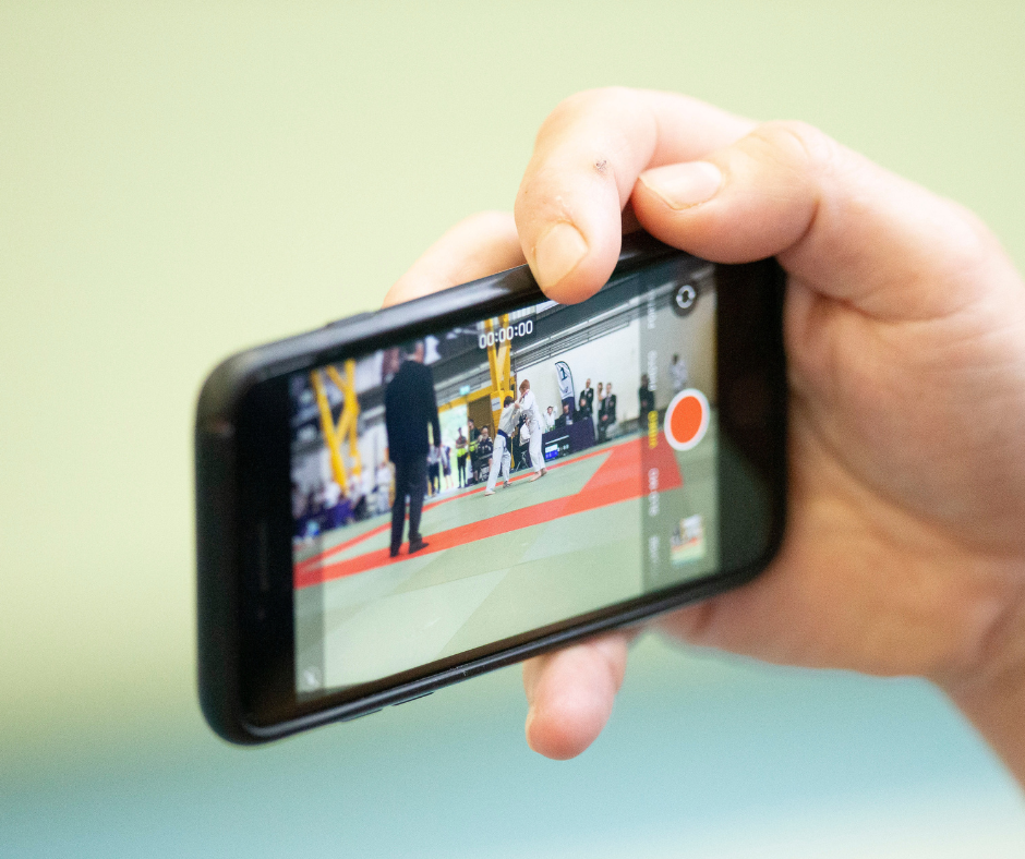 A hand holds a mobile phone that is capturing video of two children practising Judo.
