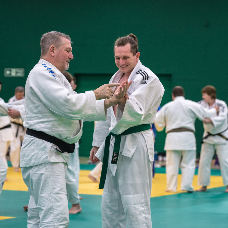 Coach and Judoka Chris Nicolstand on Mat in a busy sports hall, coach communicates with Judoka using Deafblind Dictionary.
