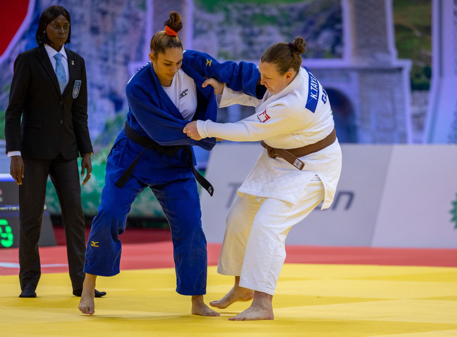 Kirsten Taylor is in a white suite gripping her opponent in blue on the tatami.