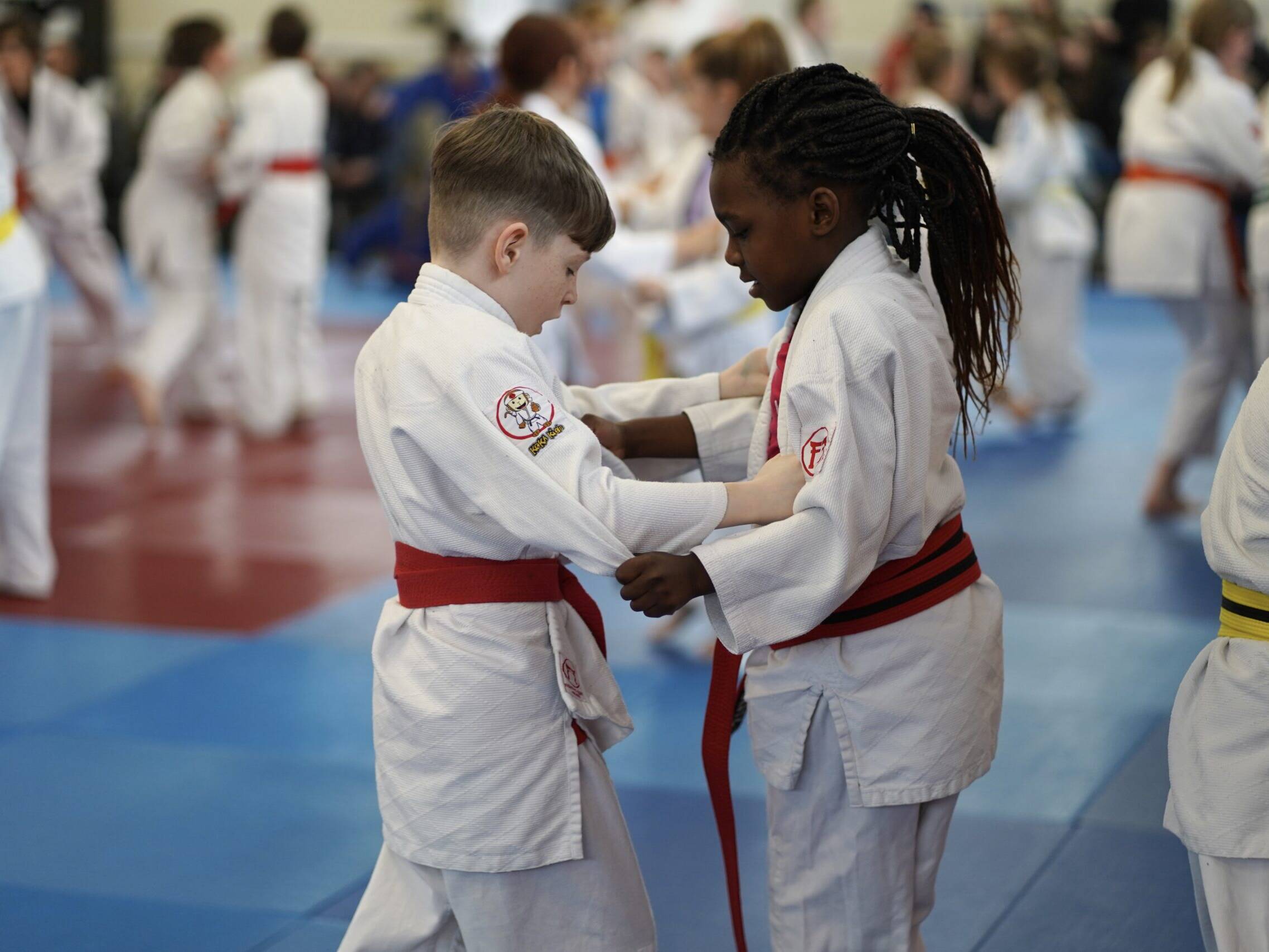 Two young judoka stand face to face gripped on at a busy club session.
