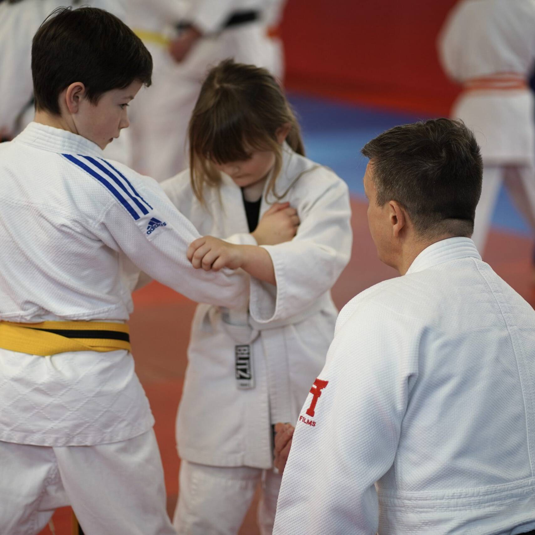 Coach kneels next to two young judoka who are gripped on to give instruction.